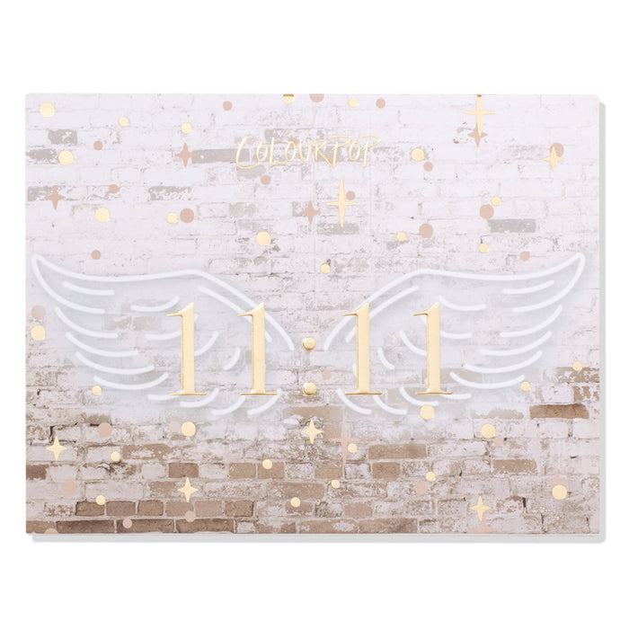 11:11 ColourPop Shadow Palette closed, featuring gold 11:11 centered between white angel wings on a faded white brick backdrop with scattered gold stars and dots, ColourPop logo above - front cover of the eyeshadow palette.
