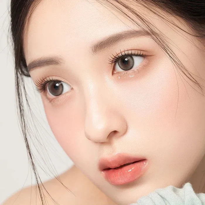 A young woman with smooth, fair skin and dewy makeup wears Gemhour Demeter 1-Day 13.0 Pebble Grey contact lenses, her light brown eyes beautifully enhanced as she gazes to the side, lips soft pink and hair pulled back.