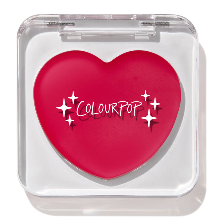 Heart-shaped hot pink compact with white “ColourPop” logo and sparkles rests closed in a clear square case, featuring the ColourPop Lip and Cheek Balm in Girls Night, a rosy multitasking balm for lips and cheeks.