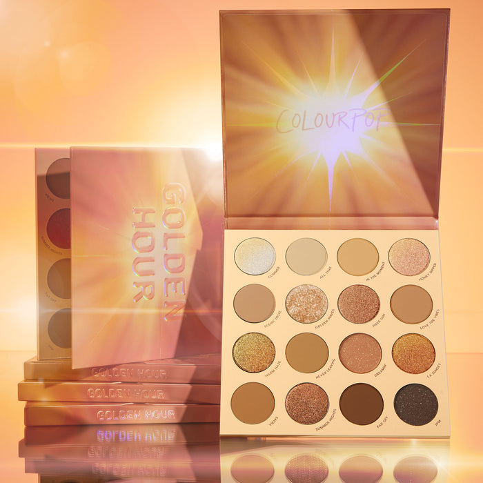 Golden Hour ColourPop Shadow Palette displaying a 16-pan mix of warm mattes, metallics, and glitter, lid with sunburst art propped open, with stacked palettes beside it on a reflective surface in a glowing golden studio.