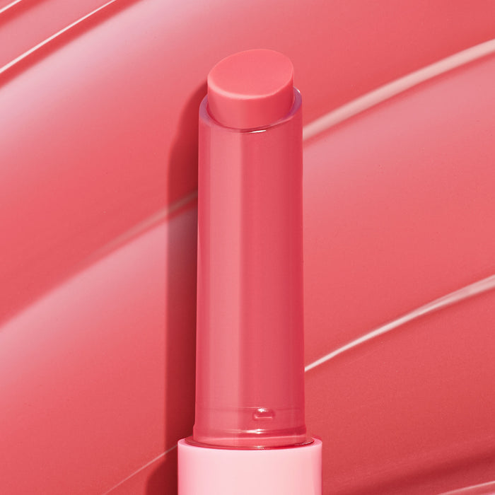 Guava Glaze shade of the ColourPop So Juicy Plumping Gloss Balm with Peptides, a twist‑up coral‑pink balm standing upright with the bullet extended, showing a glossy finish against a matching guava‑pink swirled backdrop.