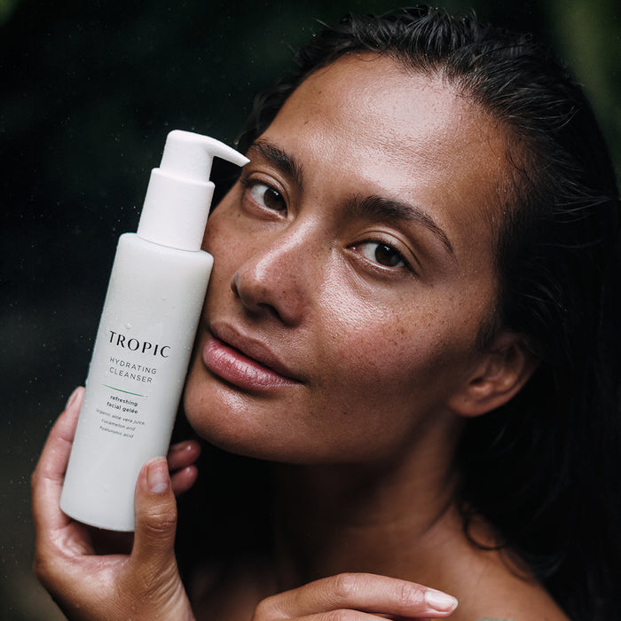 Close-up of a woman with dewy skin holding a bottle of Tropic Hydrating Cleanser next to her face, showcasing fresh, natural beauty.