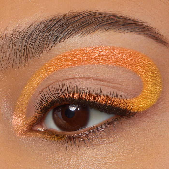 Looping graphic eyeliner tracing the crease and lower lash line in a vivid orange-to-gold chrome finish on a macro eye, showcasing Movie Star, the ColourPop Chrome Crème Gel Liner, with long lashes and a groomed brow.