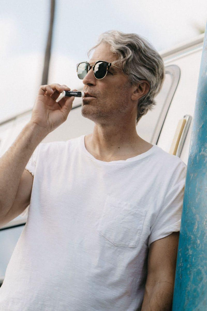 Man with gray hair and sunglasses applying chapstick while leaning against a blue pole and wearing a white t-shirt.