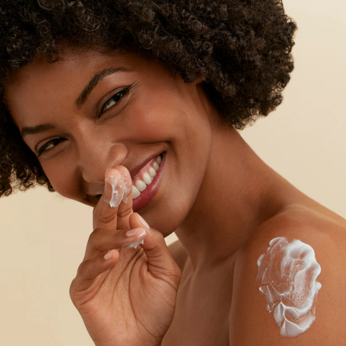 Woman applying rich white skincare cream to shoulder and fingers, showcasing texture for all skin types