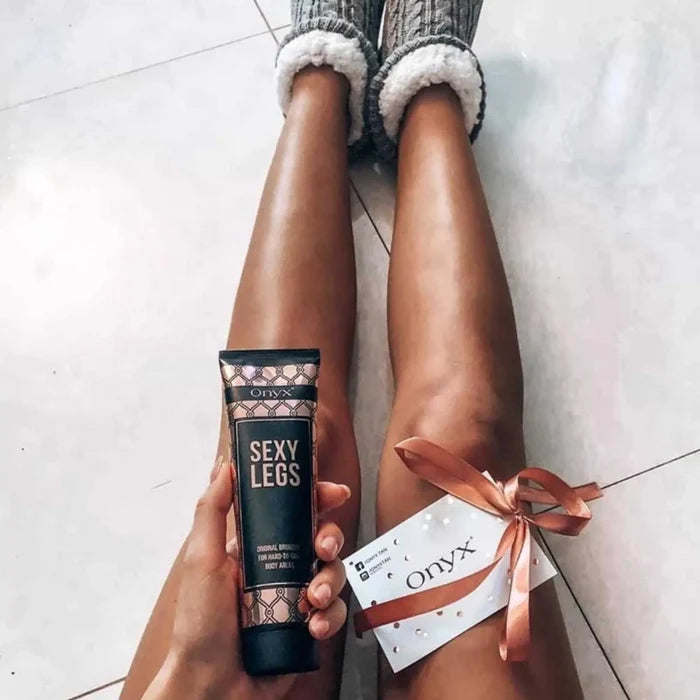 Kobieta siedzi na podłodze z wyprostowanymi nogami, trzyma balsam Onyx Sexy Legs i kartkę z logo marki przewiązaną wstążką