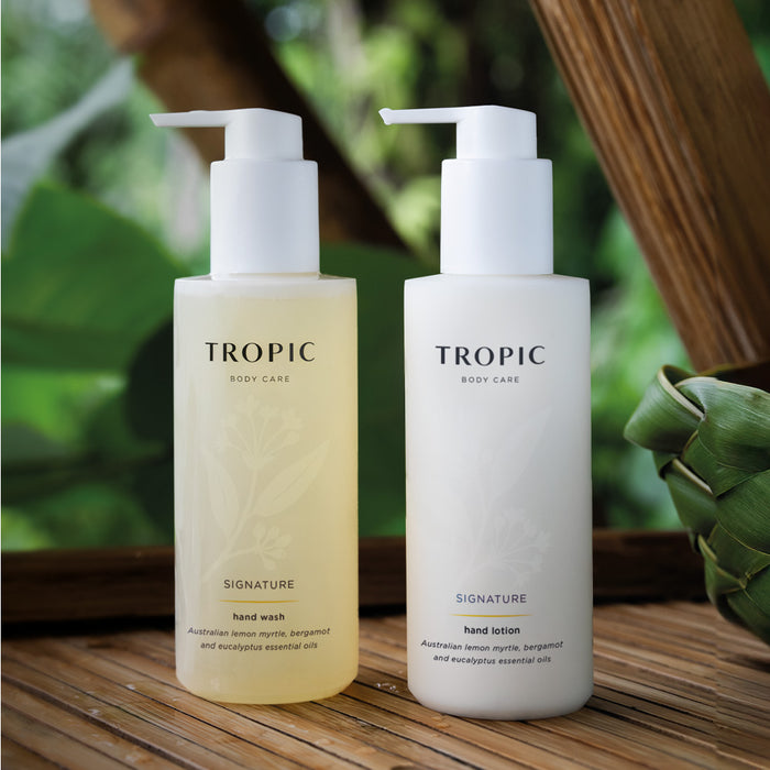 Image shows Tropic Body care Signature hand wash and Signature hand lotion bottles with white  dispensing pumps. There is a faint floral design on the bottle. The products contain Australian lemon myrtle, bergamot and eucalyptus essential oils. The bottles are placed on a wooden surface with green leaves in the background.