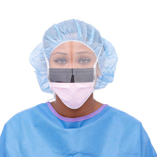 A woman, wearing a surgical cap, mask, and face shield, is looking directly at the viewer. She is dressed in light blue scrubs.