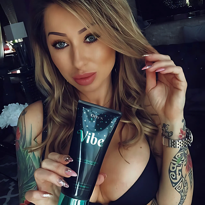 Kobieta z tatuażem i falowanymi włosami trzyma Onyx Vibe. Przyspieszacz do opalania wspiera naturalny koloryt i pielęgnuje skórę.